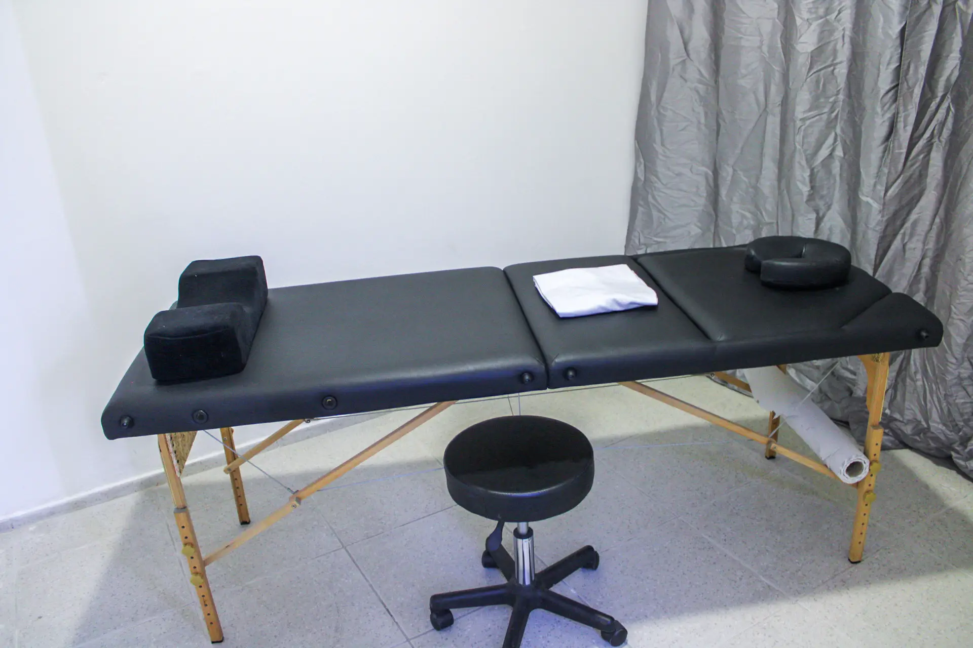 Sala de osteopatia com marquesa preta, almofadas, toalha e banco, junto a uma cortina cinzenta
