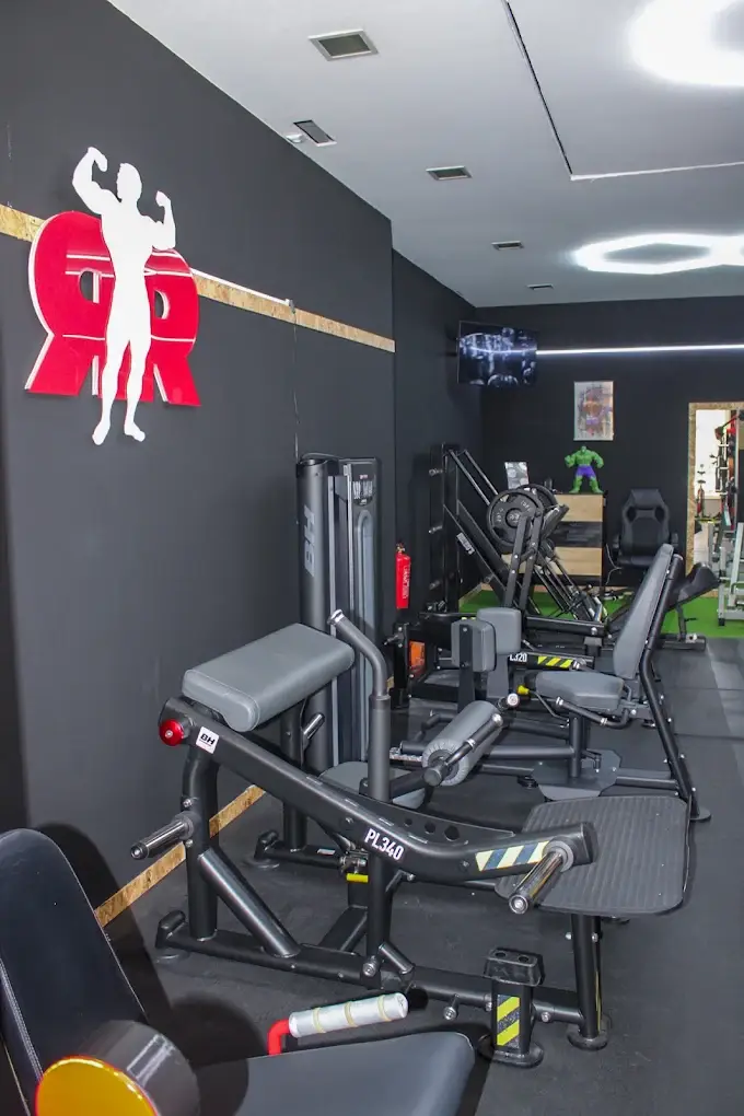 Sala de treino com paredes pretas, faixa de madeira, monograma RR e máquinas de pernas alinhadas