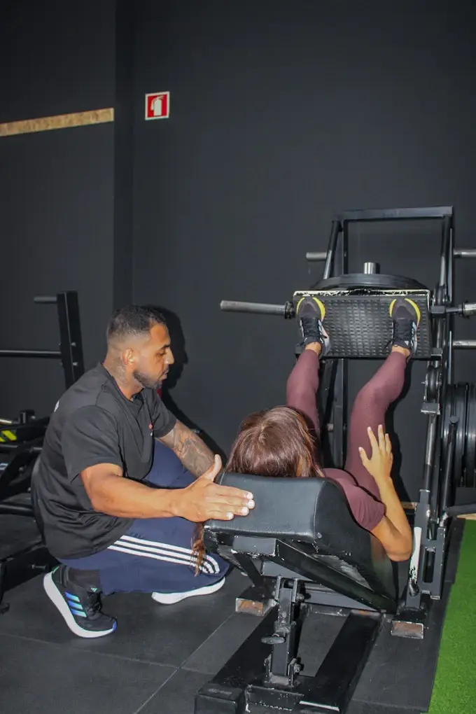 Ruan Ribeiro ajoelhado ao lado de uma cliente, a acompanhar um exercício de leg press